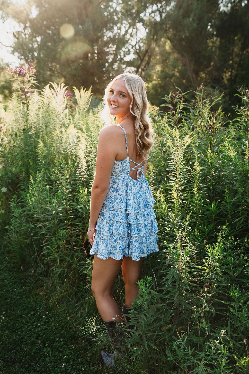 A person in a blue floral dress poses in a garden with tall green plants during golden hour sunlight.