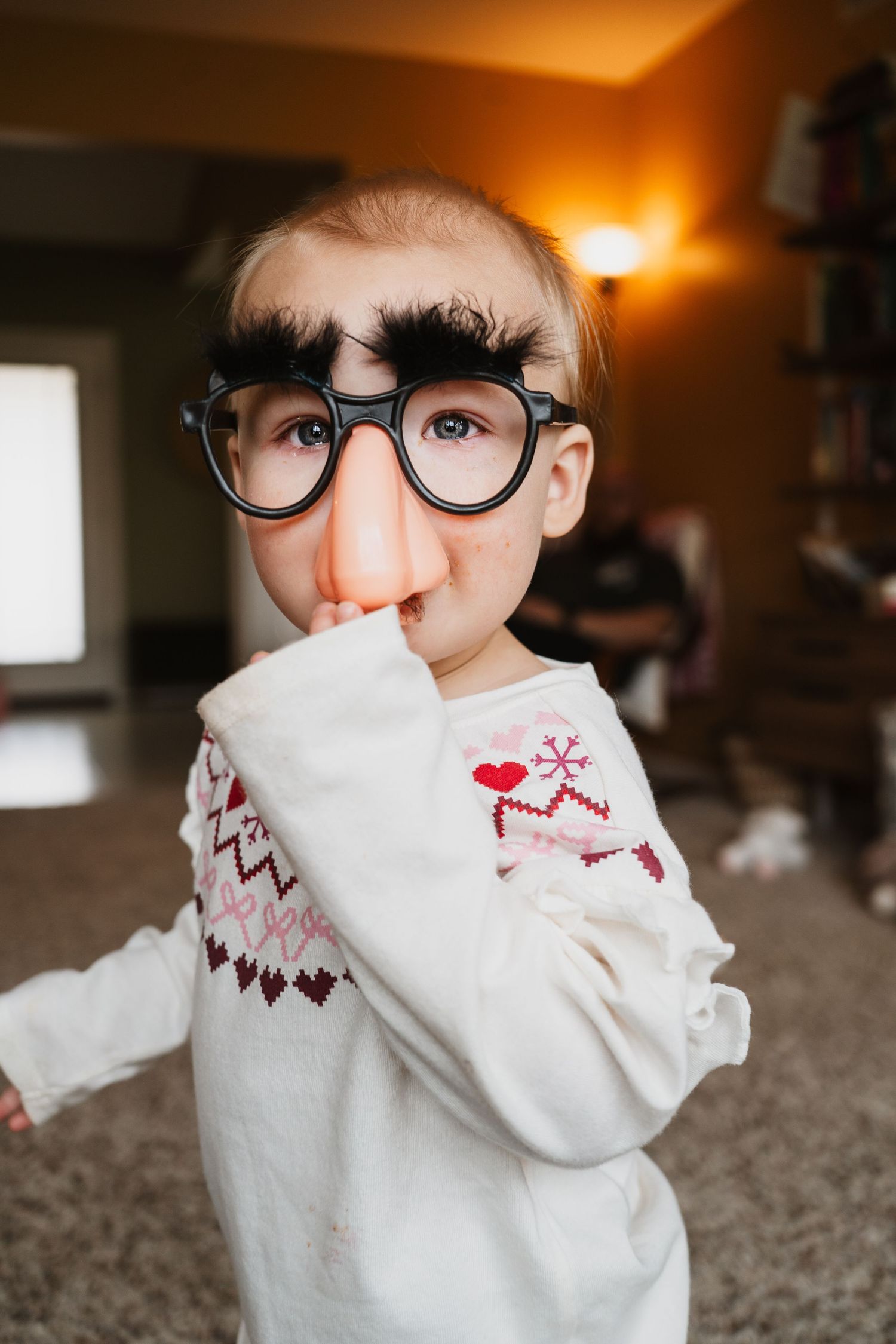 Young child wearing funny Groucho Marx disguise glasses with big nose and bushy eyebrows indoors.