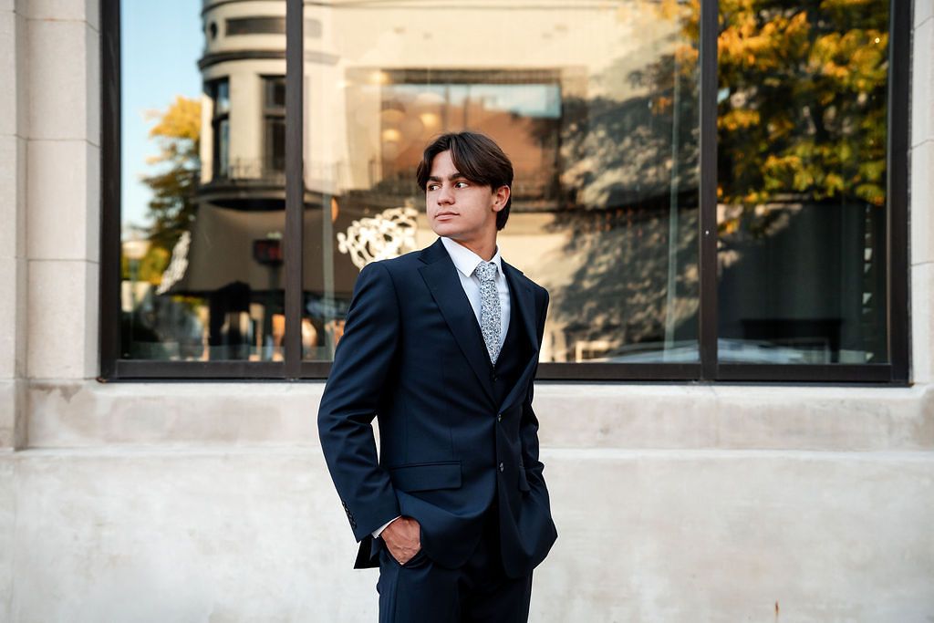 Young man in navy suit with tie poses confidently outdoors near modern building with glass windows.