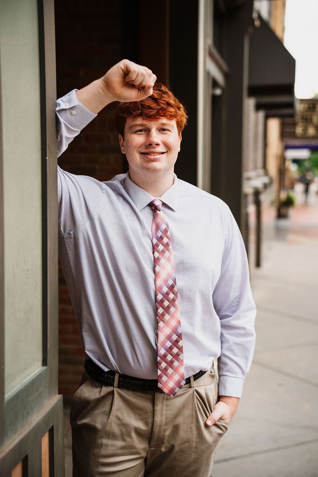 Young man with red hair wearing a plaid tie and dress shirt, leaning against a building on a city sidewalk.