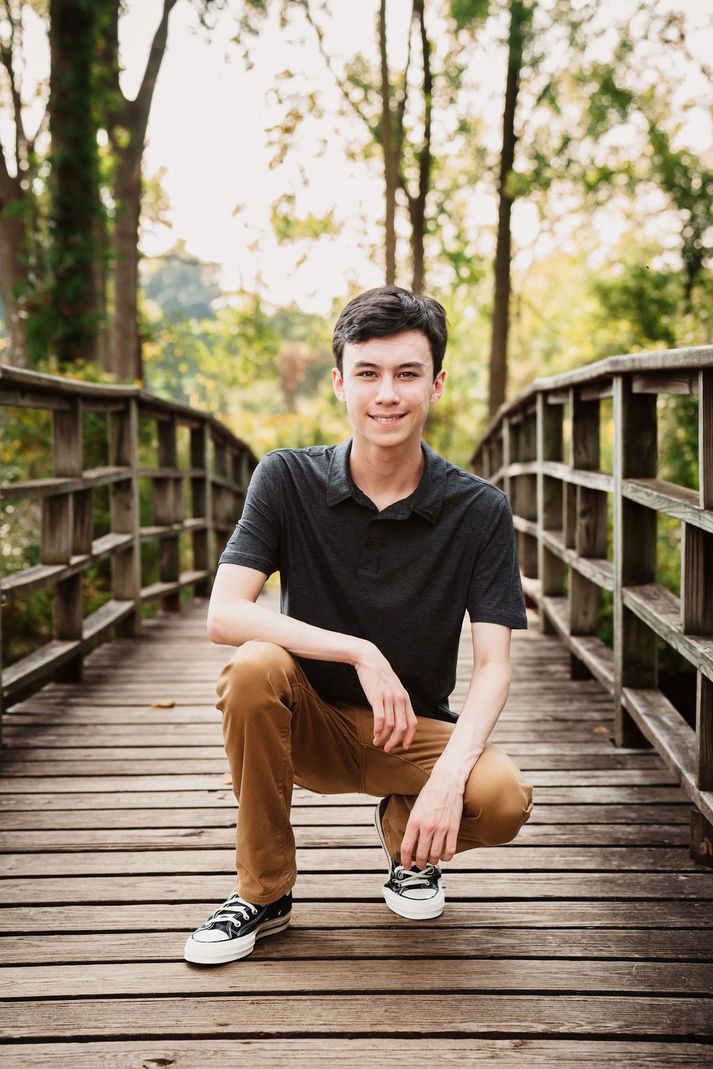 Young man in black polo and tan pants crouching on a wooden bridge surrounded by lush green trees.