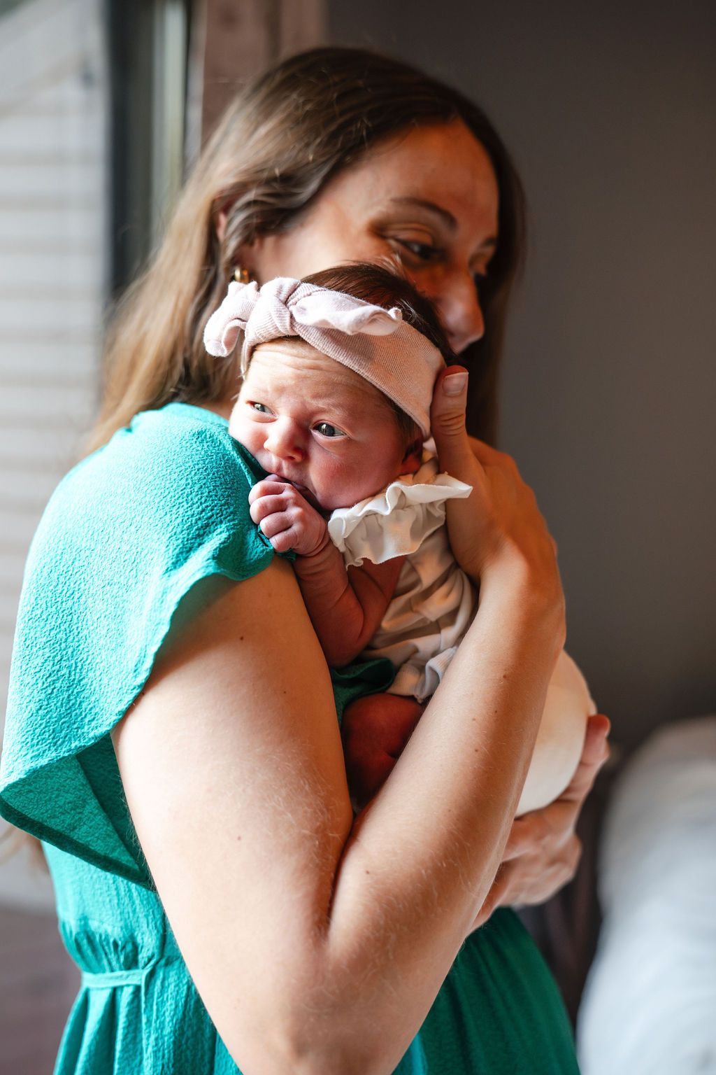 A mother in a teal dress tenderly holds her newborn baby wearing a pink bow headband near a window.