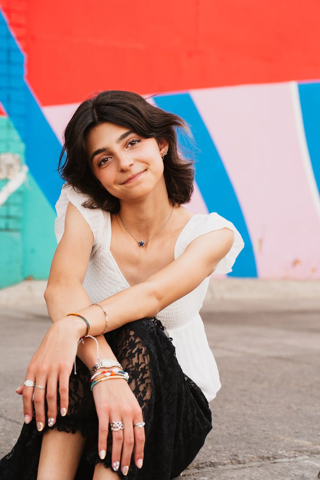 A young woman in a white top and black lace skirt sits against a colorful mural wall wearing layered bracelets and rings.