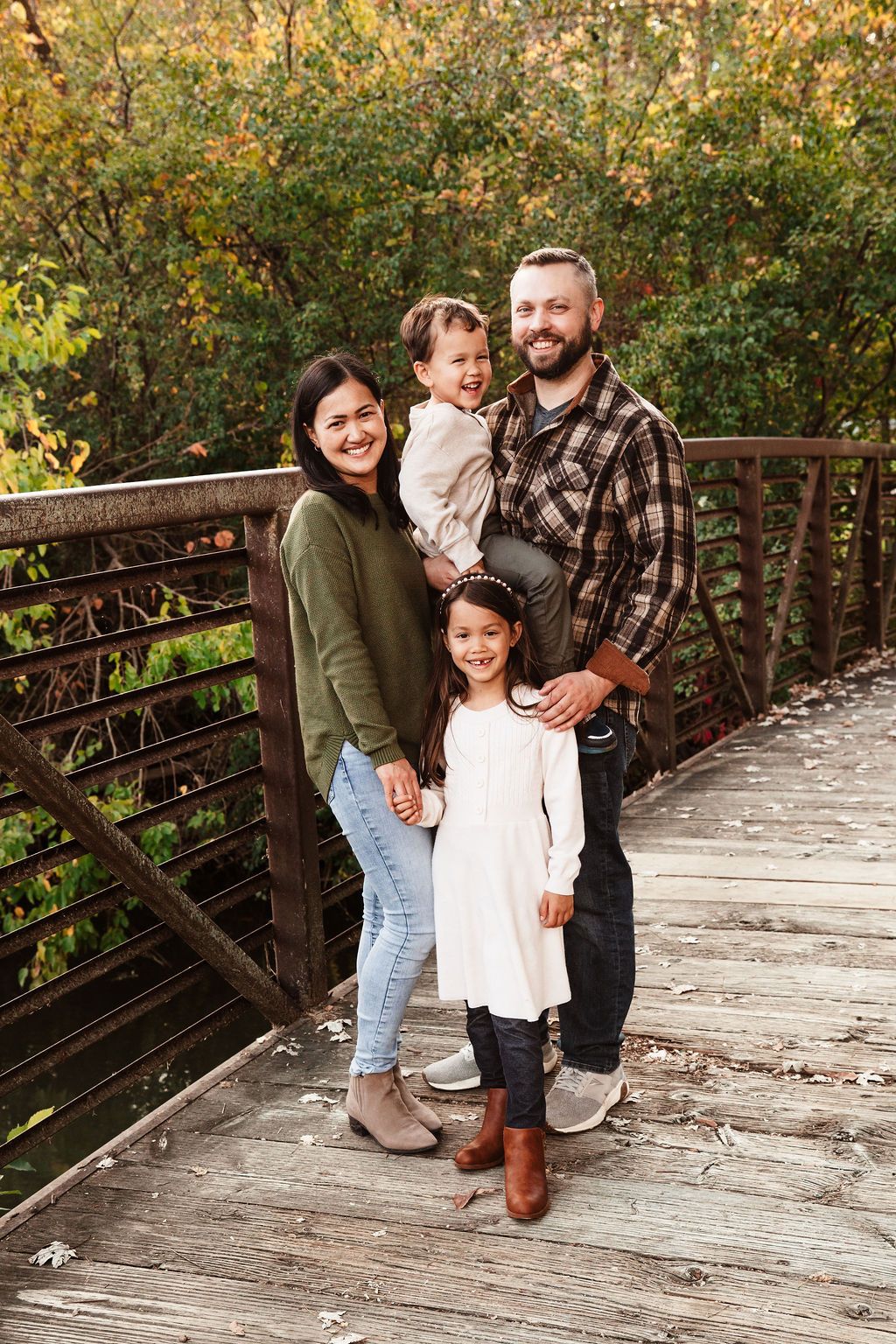 Family of four smiling on wooden bridge surrounded by lush green fall foliage, parents holding two young daughters.