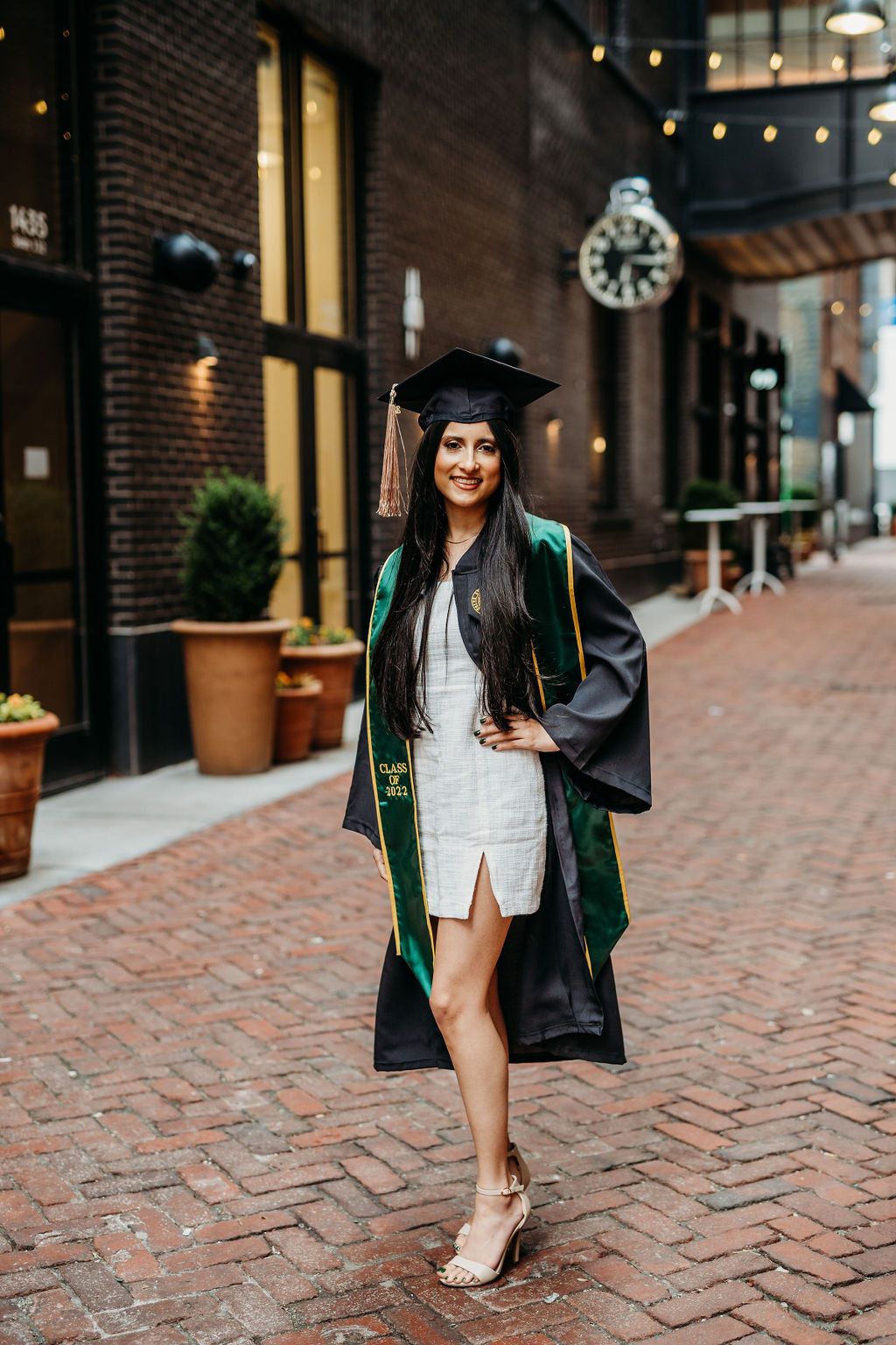 Graduate in cap and gown poses on brick walkway with urban architecture and storefronts in background.