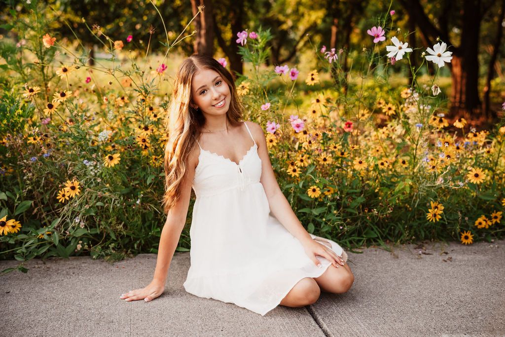 A person in a white sundress sits on a pathway surrounded by vibrant yellow wildflowers in a sunny garden setting.