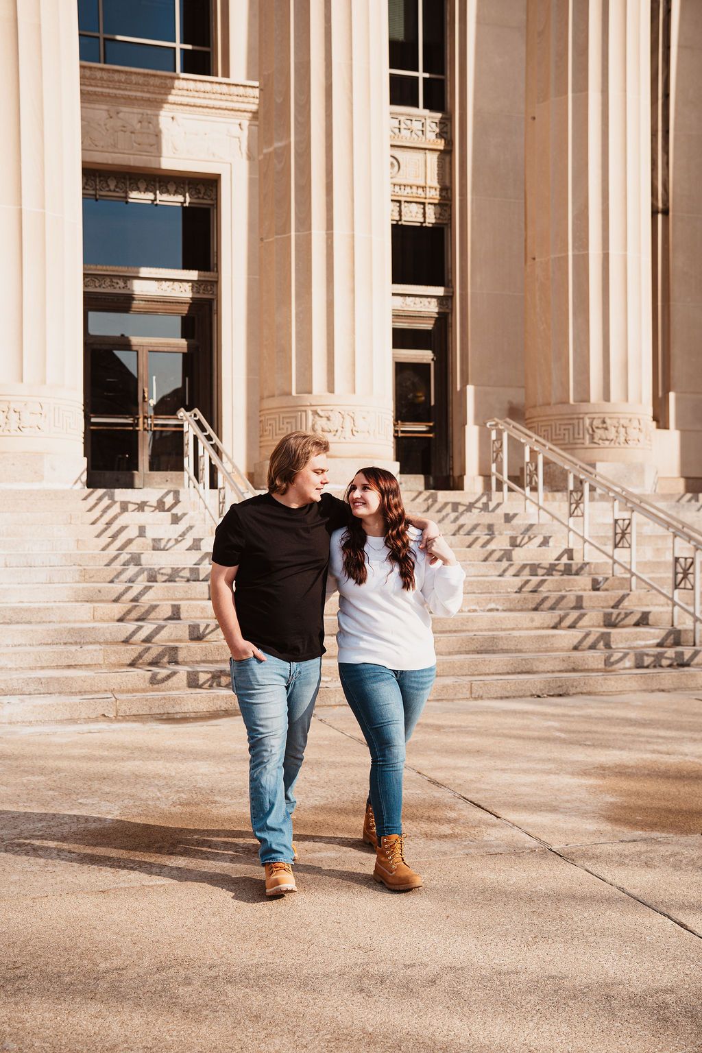 Couple walking together in front of a grand building with stone columns, wearing casual jeans and boots.