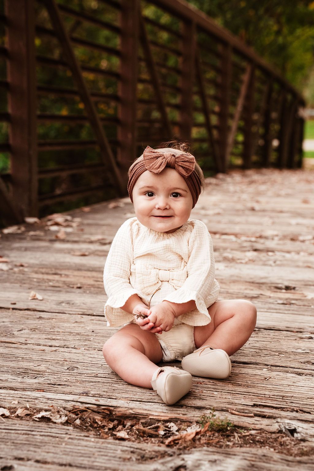 Smiling baby girl in cream outfit and bow headband sitting on wooden bridge deck surrounded by autumn leaves.