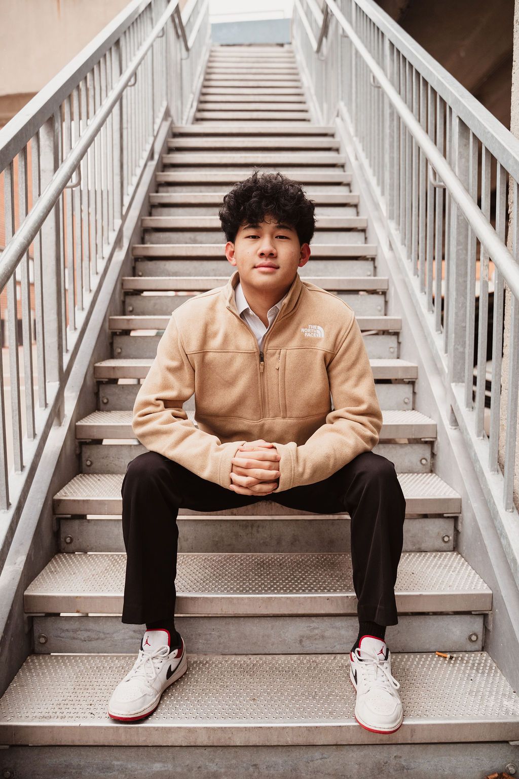 Young man in a beige North Face jacket sitting on metal stairs with hands clasped, wearing Jordan sneakers.