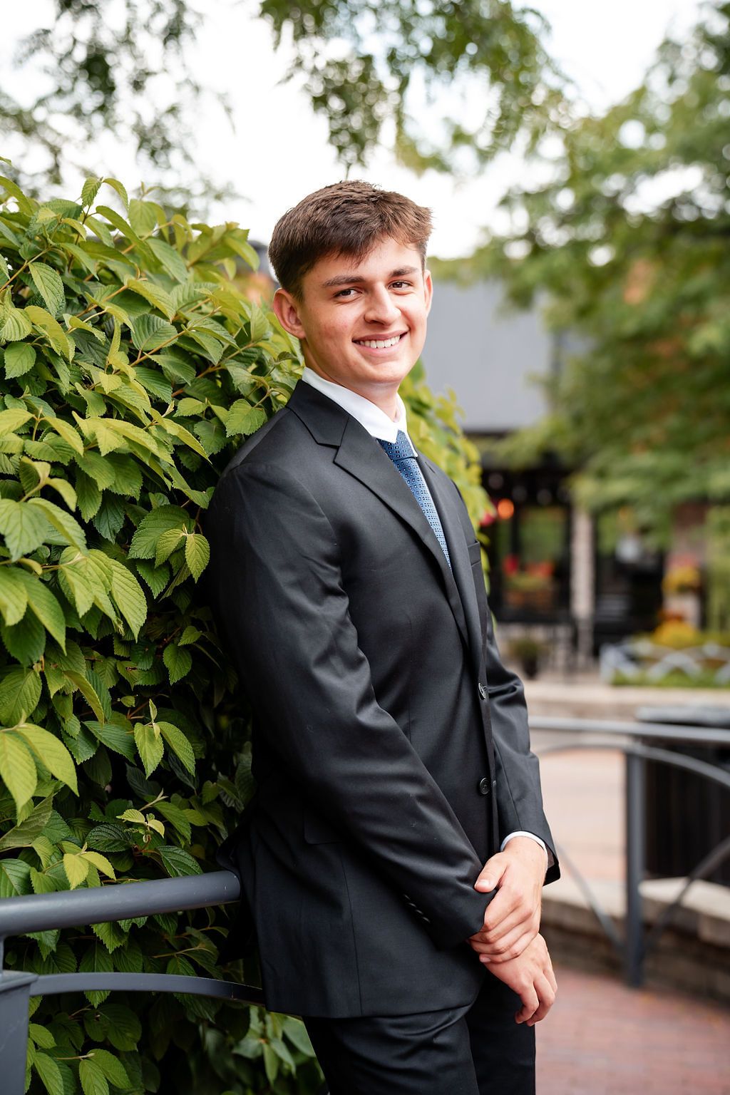 Young man in dark suit and blue tie smiling outdoors near green ivy wall.