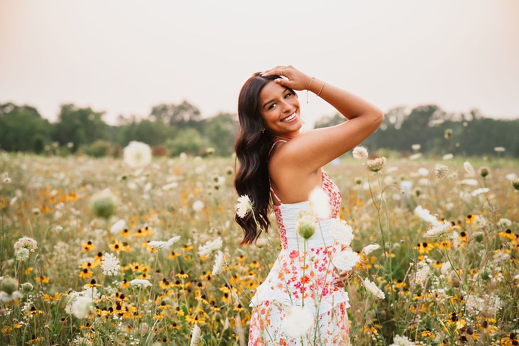 Person in white dress poses among blooming wildflowers in sunny meadow.