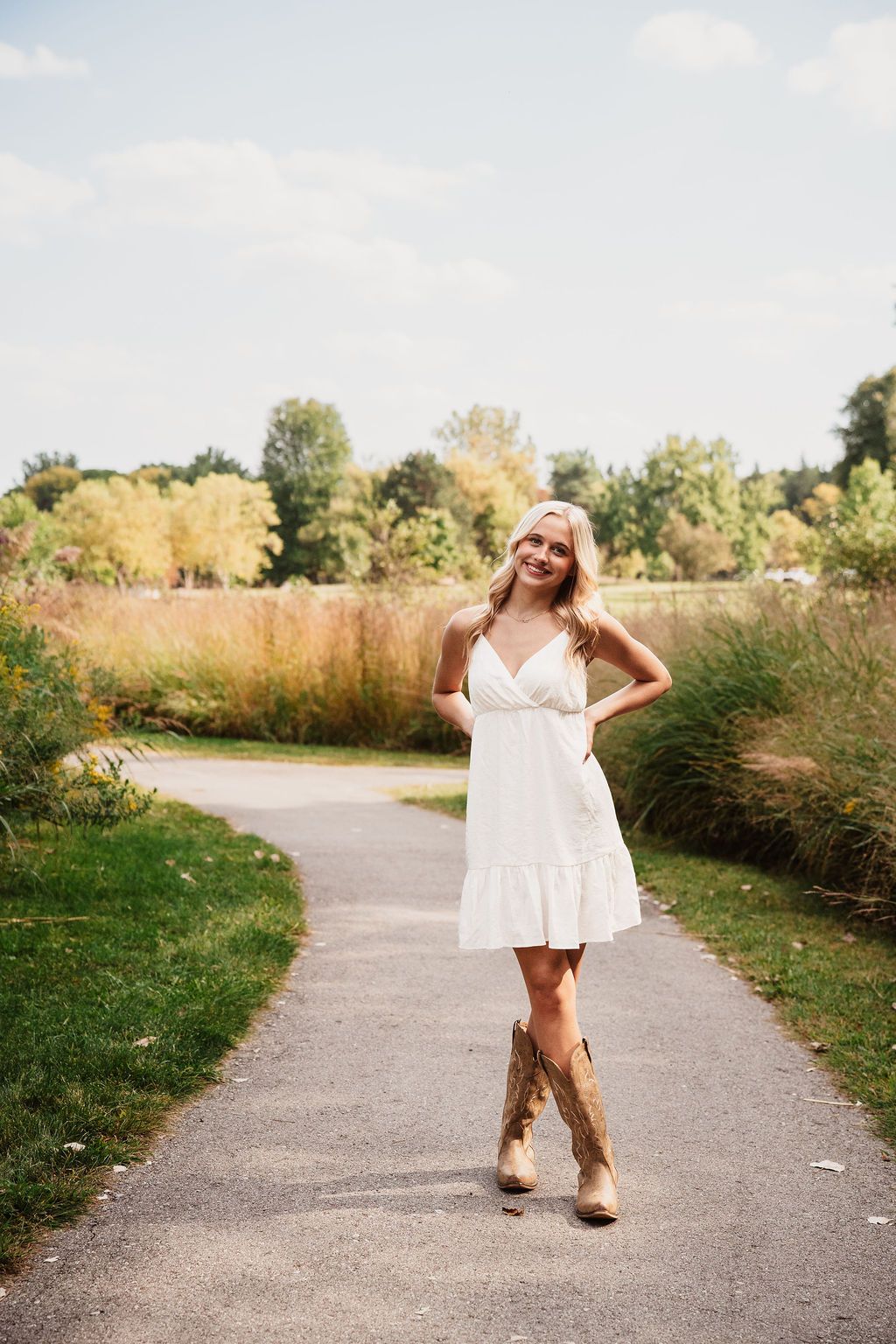 Smiling blonde woman in white sundress and cowboy boots standing on a winding path in a lush green park.