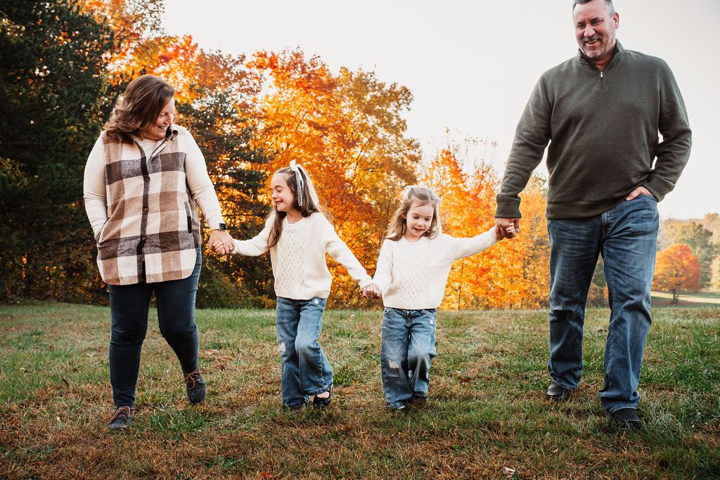 Family of four holding hands in a sunny autumn field surrounded by colorful fall foliage trees.