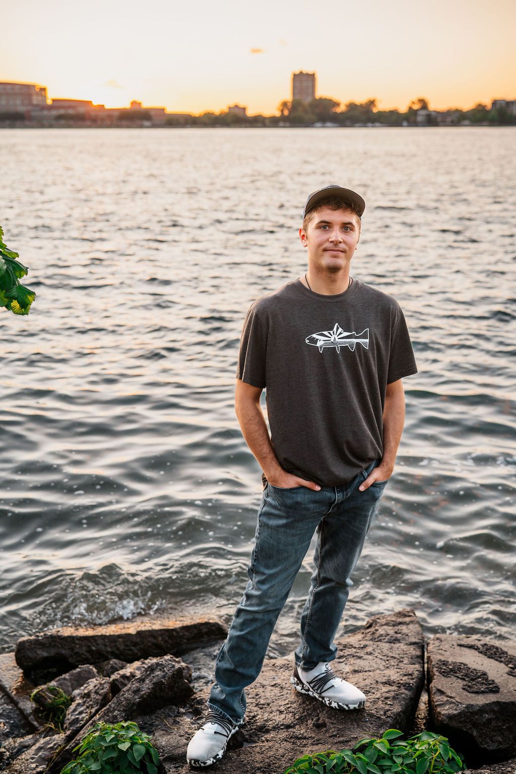 Young man in gray t-shirt and jeans stands on rocky shoreline at sunset with city skyline reflected on calm water.