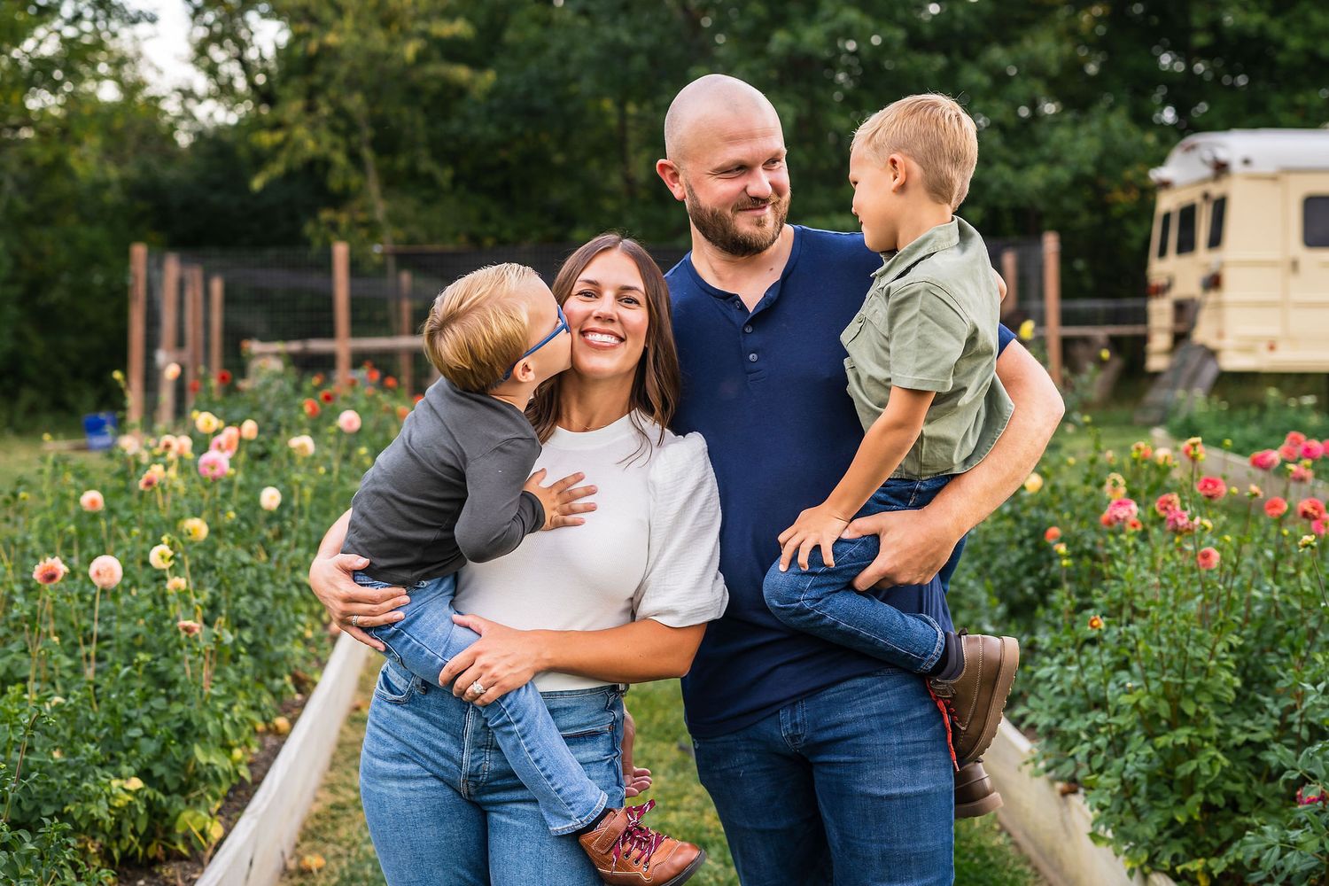Happy family of four smiling outdoors in a flower garden, parents holding two young boys in a warm, candid portrait.