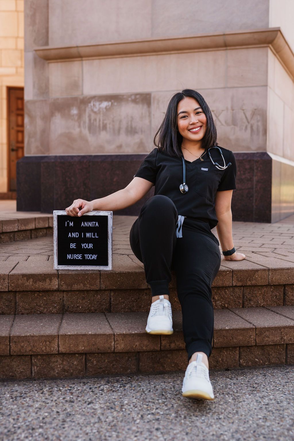 Healthcare worker in black scrubs and white sneakers sits on stone steps holding a letter board sign outdoors.