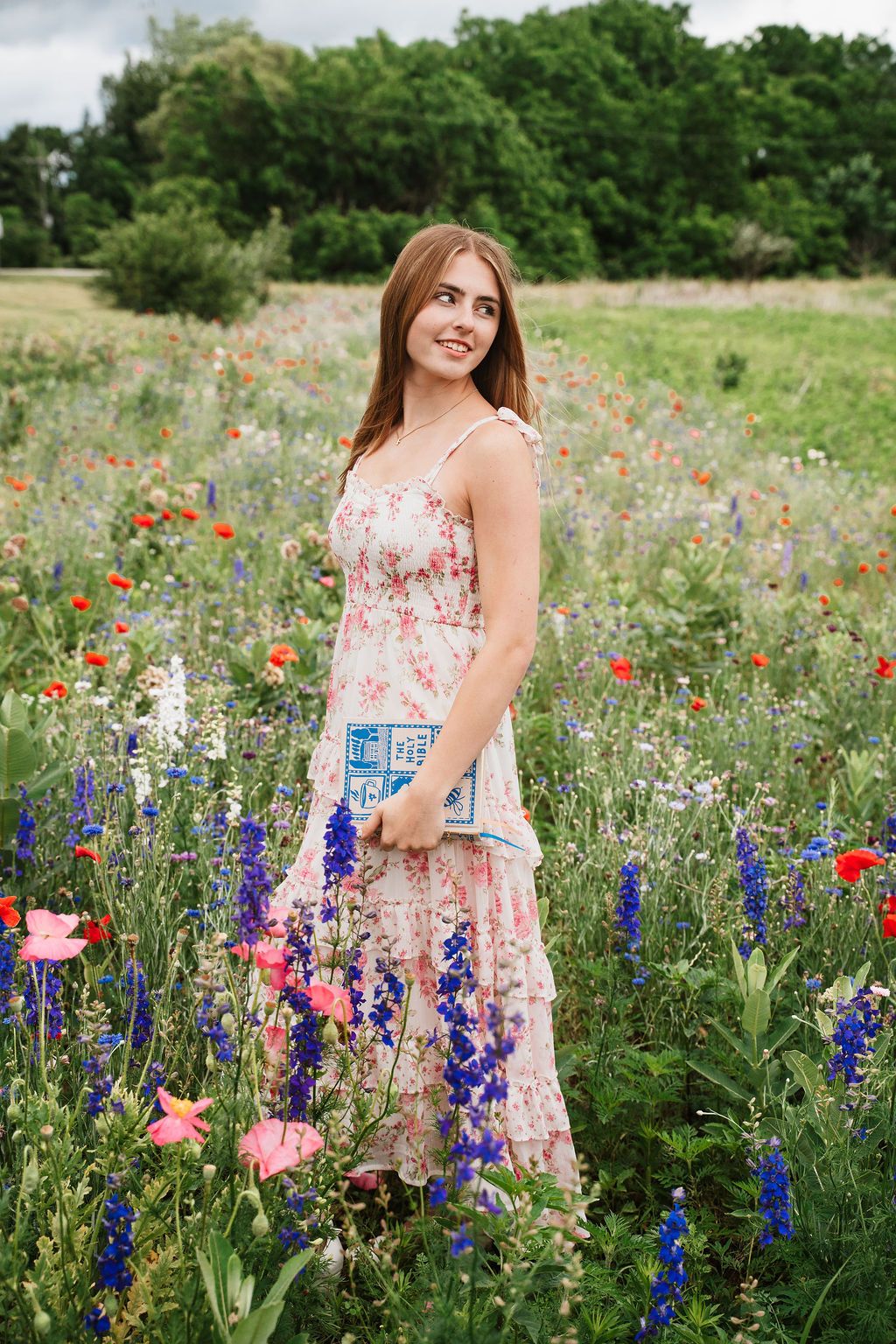 Young woman in floral maxi dress standing in colorful wildflower field with poppies and blue delphiniums.