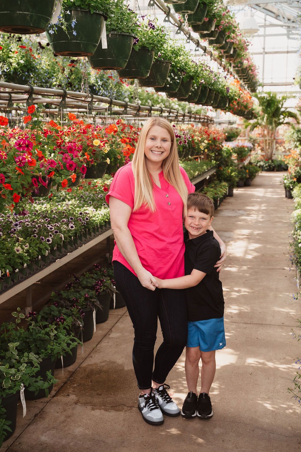 A smiling woman in a pink shirt hugs a young boy in a black shirt inside a colorful flower greenhouse.