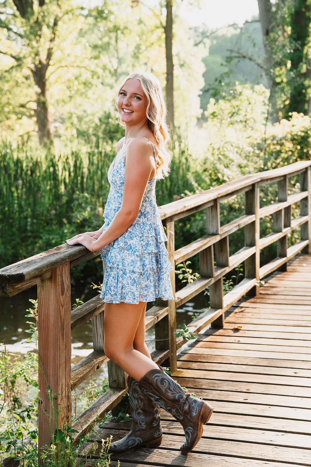 Young blonde woman in floral mini dress and cowboy boots smiling on a rustic wooden bridge surrounded by lush greenery.