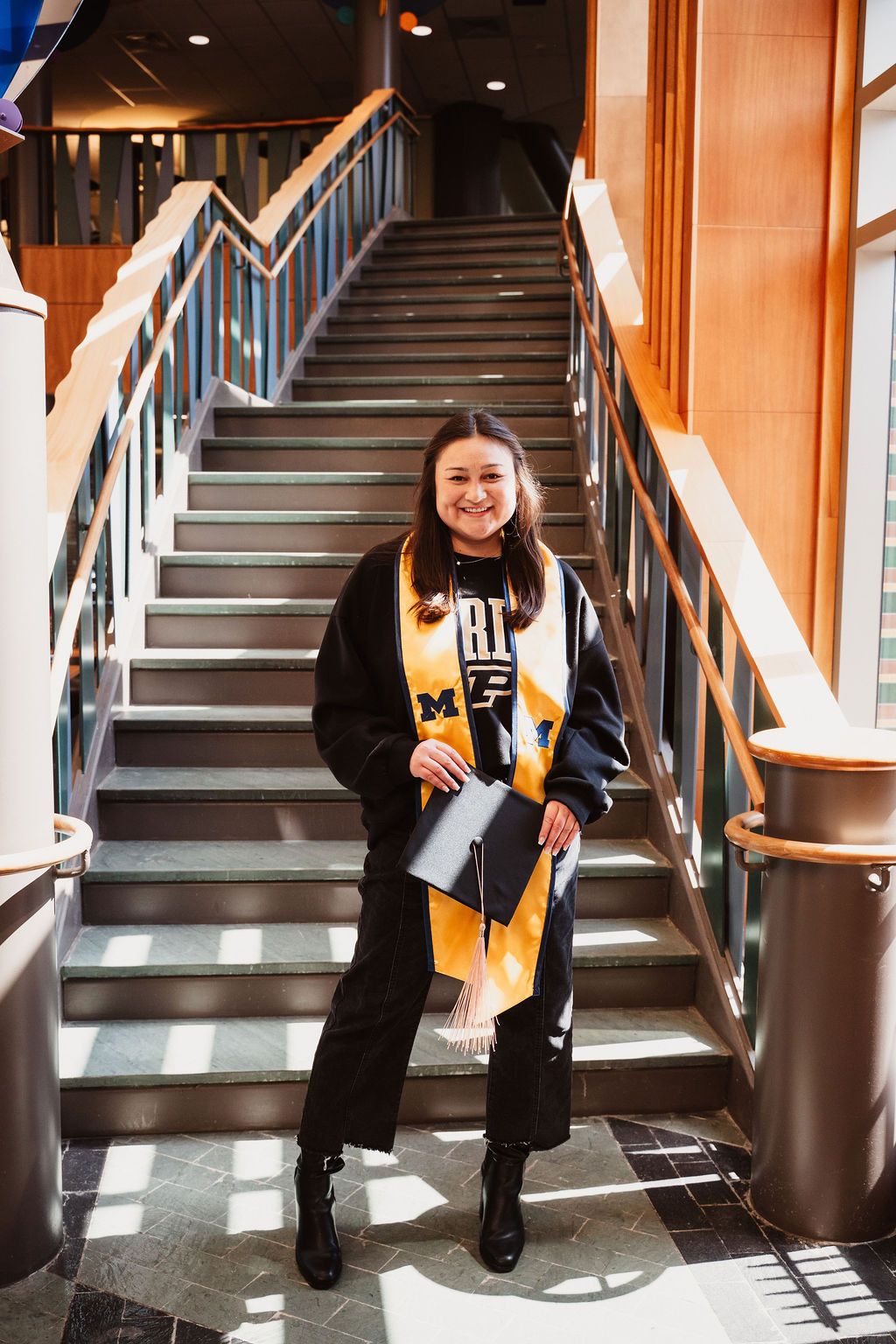 Graduate wearing Michigan stole holding cap while posing on modern interior staircase.