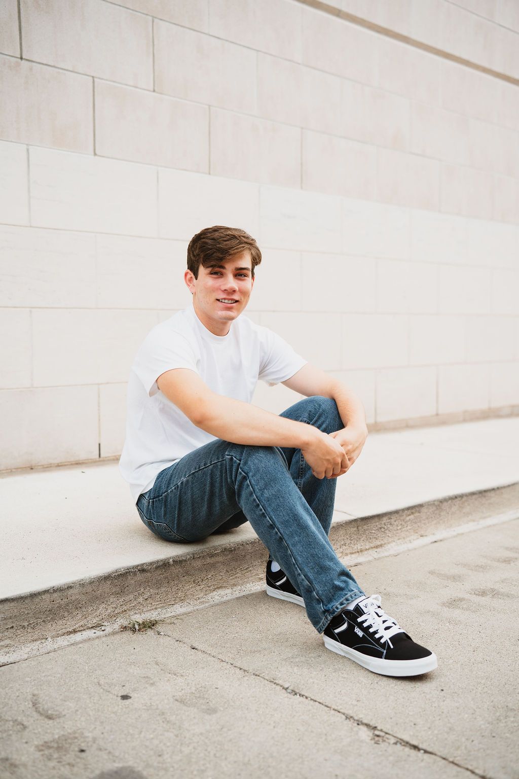 Young man in white t-shirt and jeans sitting on steps against a light stone wall, smiling at camera.