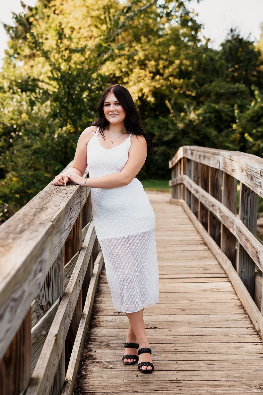 Young woman in white midi dress and black heels posing on a wooden bridge surrounded by lush green trees.