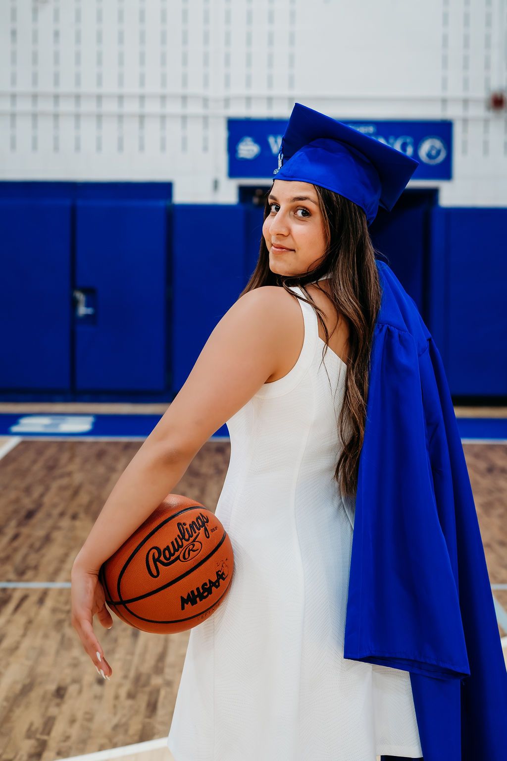 Graduate in white dress holding basketball and wearing blue cap and gown poses in gymnasium.