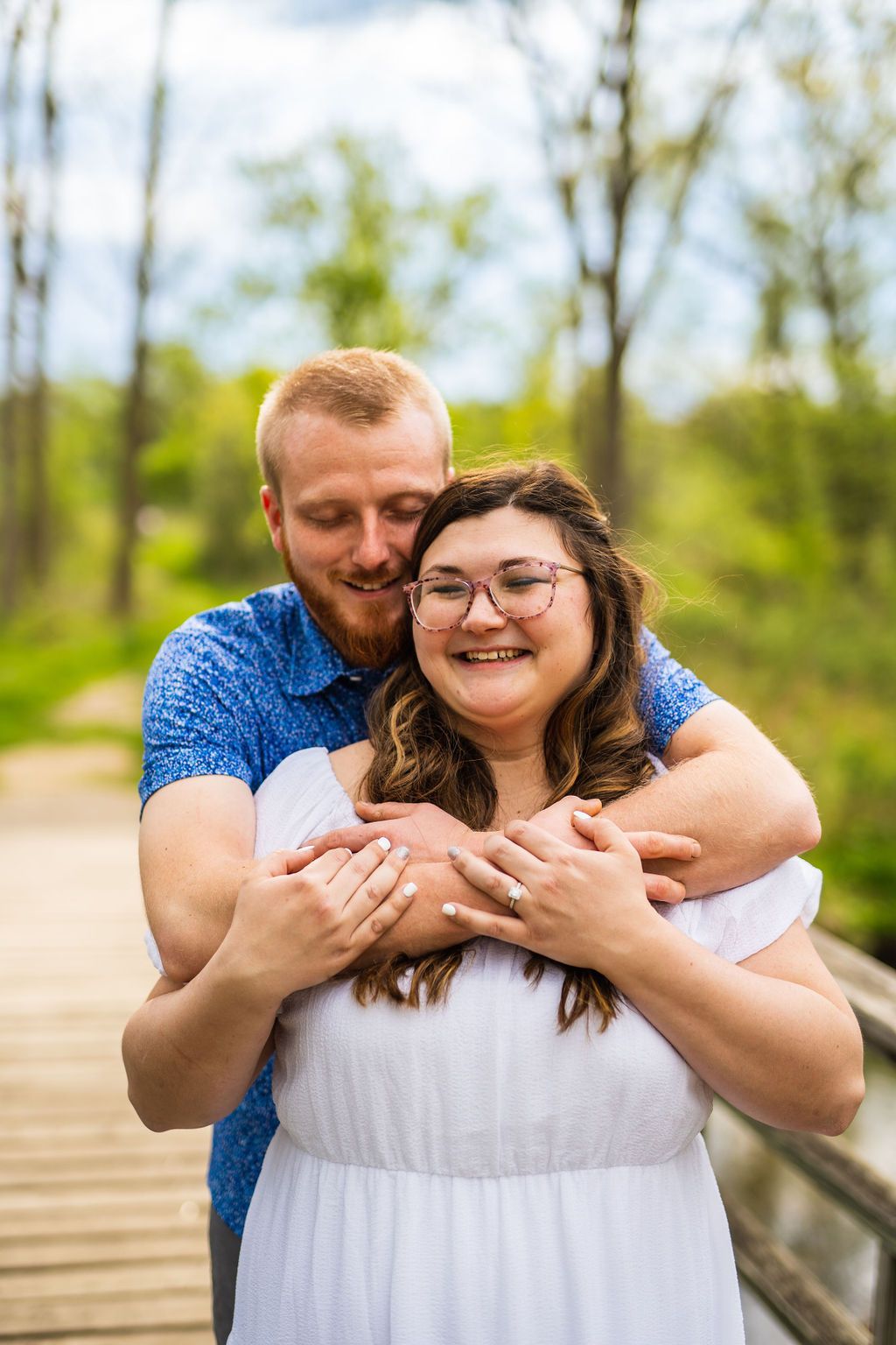 A couple embraces on a wooden bridge surrounded by lush greenery on a bright sunny day.