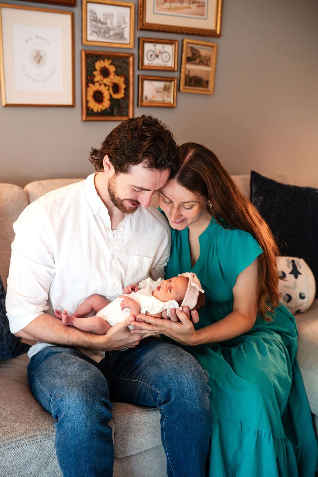 A couple sits on a couch, tenderly holding their newborn baby, with a gallery wall of framed art behind them.