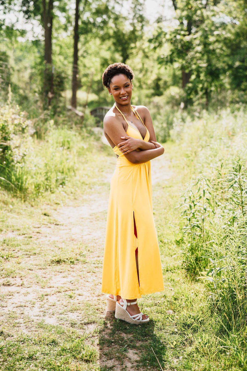 A smiling woman in a yellow halter maxi dress with a front slit stands on a garden path surrounded by lush greenery.