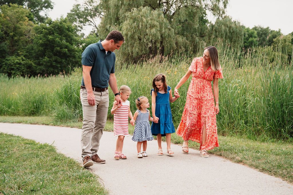 Family of five walking on a park path, parents with three young girls in colorful dresses enjoying a sunny outdoor stroll.