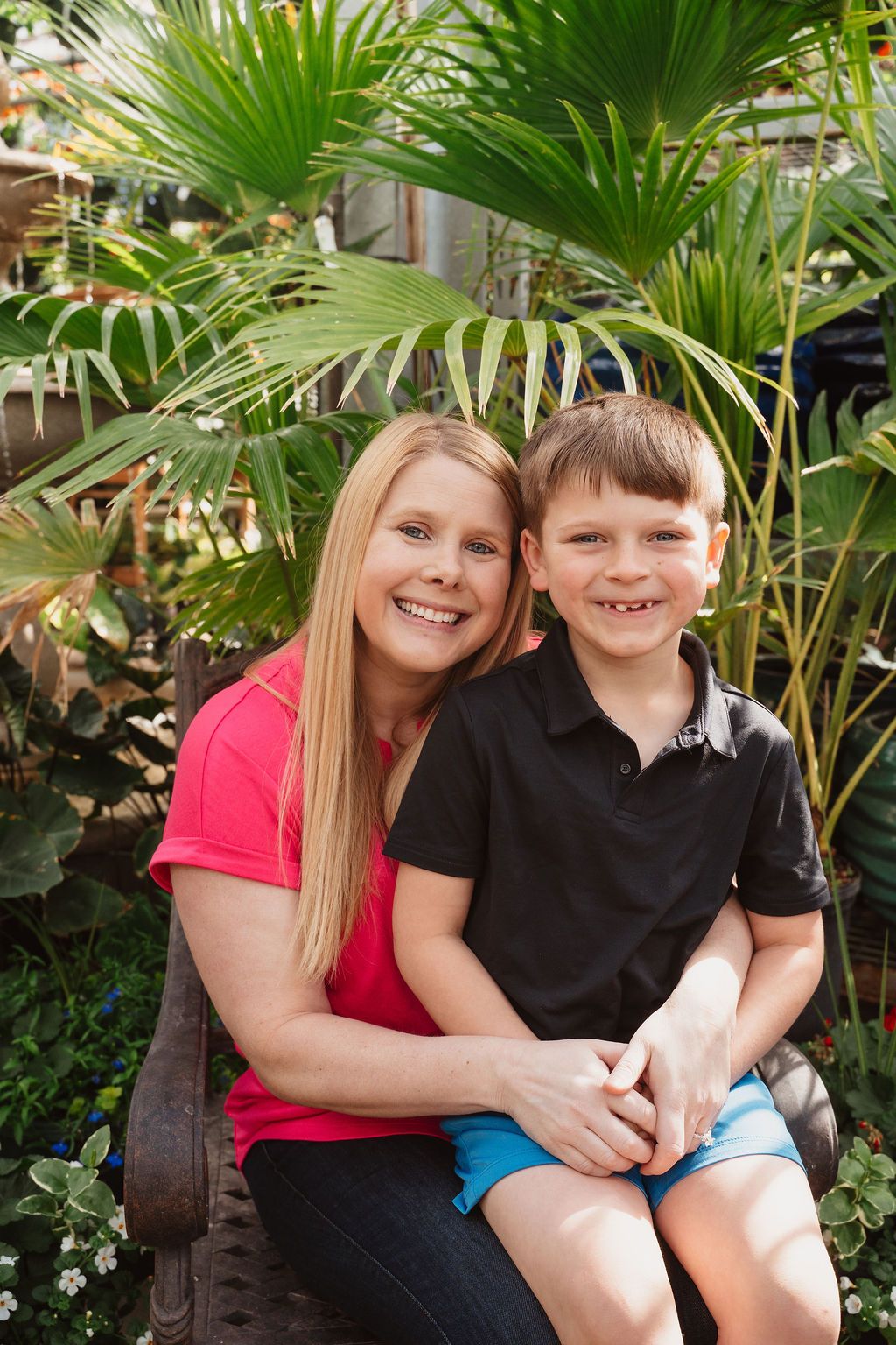 A smiling woman and young boy pose together on a bench surrounded by tropical palm plants.