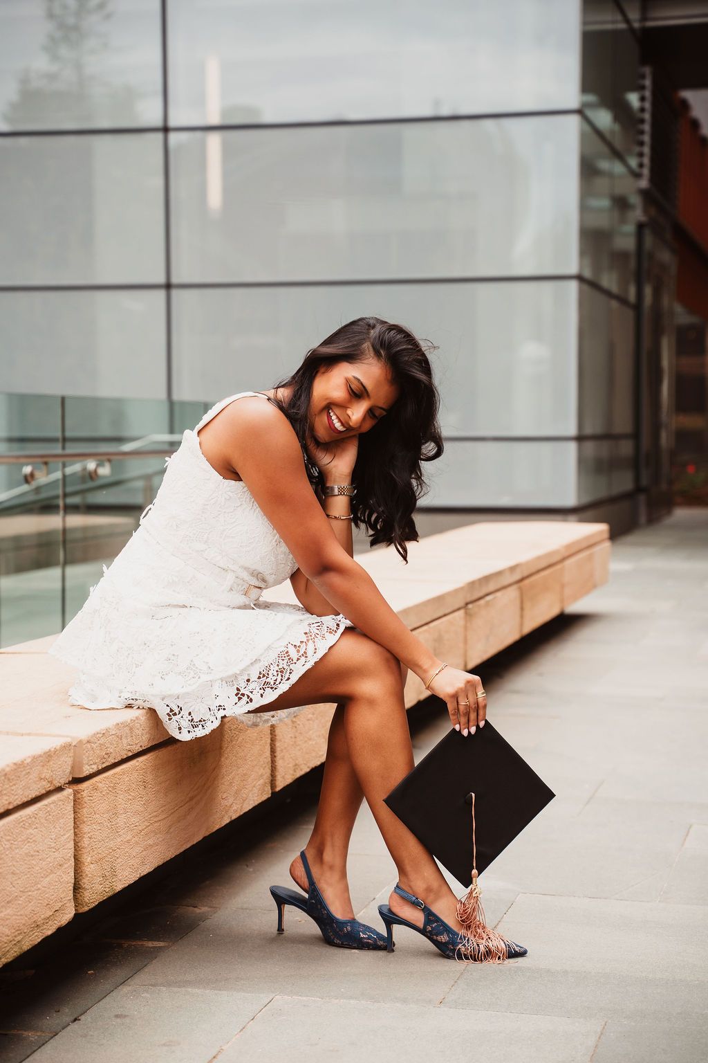 Smiling graduate in white lace dress holding cap, sitting on bench outside modern building wearing blue heels.