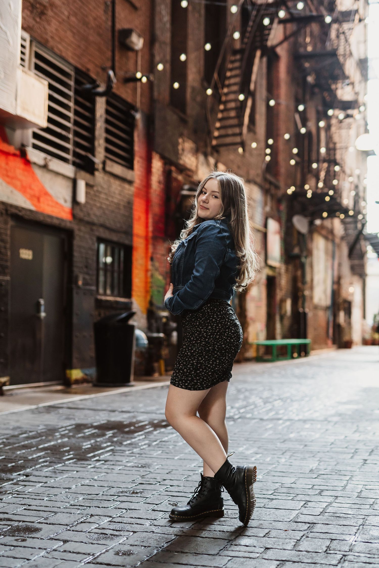 Young woman in denim jacket, floral mini skirt, and black boots posing in a brick alley with string lights.