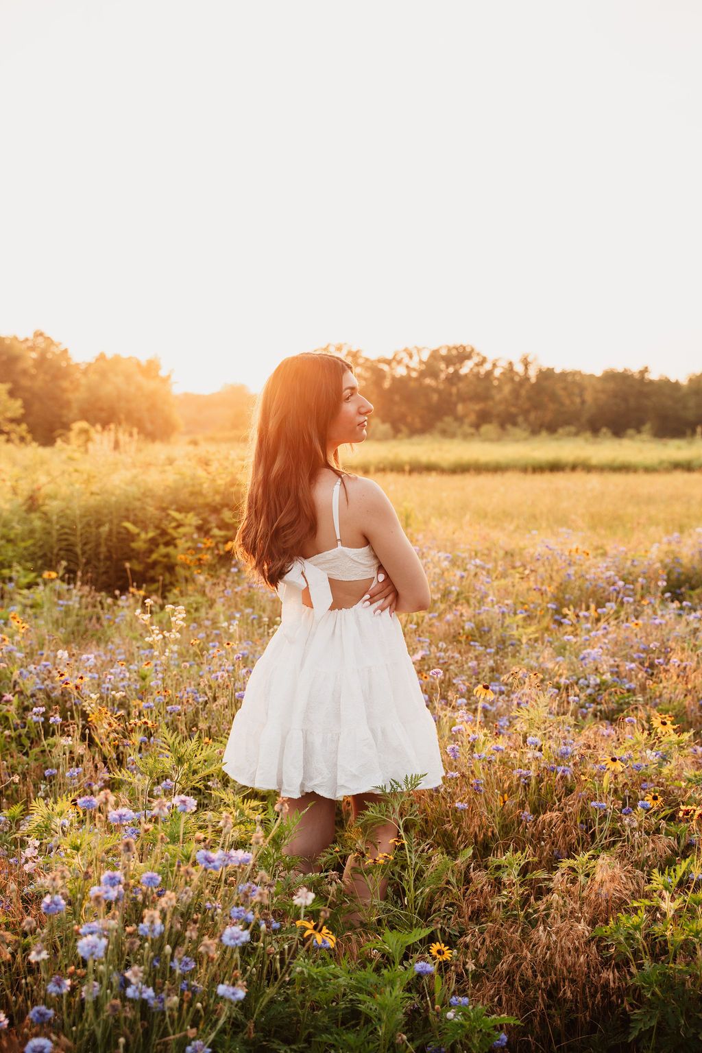 Young woman in white sundress standing in wildflower field at golden sunset, smiling over shoulder.