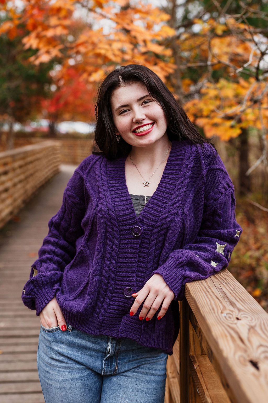 A person in a purple knit cardigan poses on a wooden bridge against vibrant autumn foliage in the background.