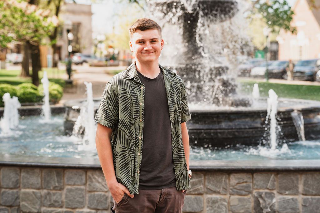 Young man smiling in green floral shirt standing in front of a decorative fountain in an outdoor plaza.