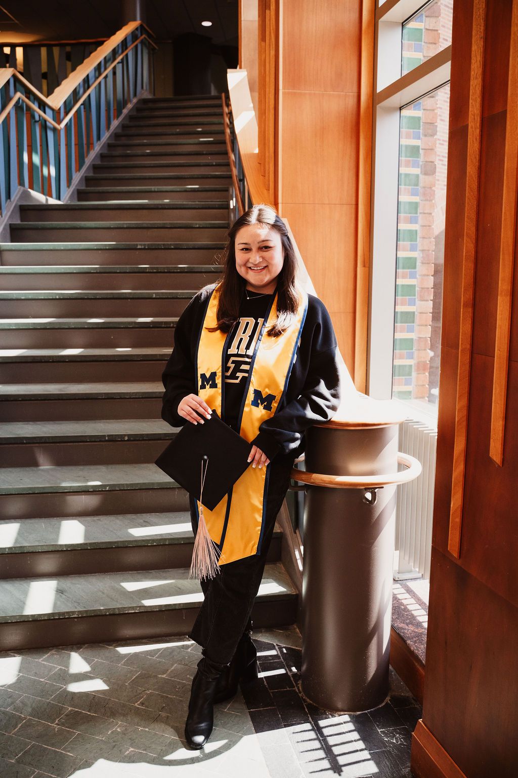 Smiling graduate in black gown and gold Michigan stole poses by staircase holding cap indoors.