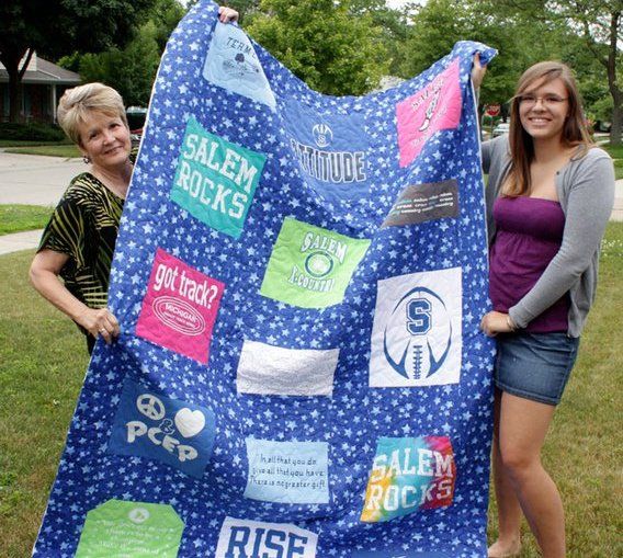 Two women proudly display a blue star-print memory quilt featuring Salem Rocks and other colorful T-shirt panels outdoors.