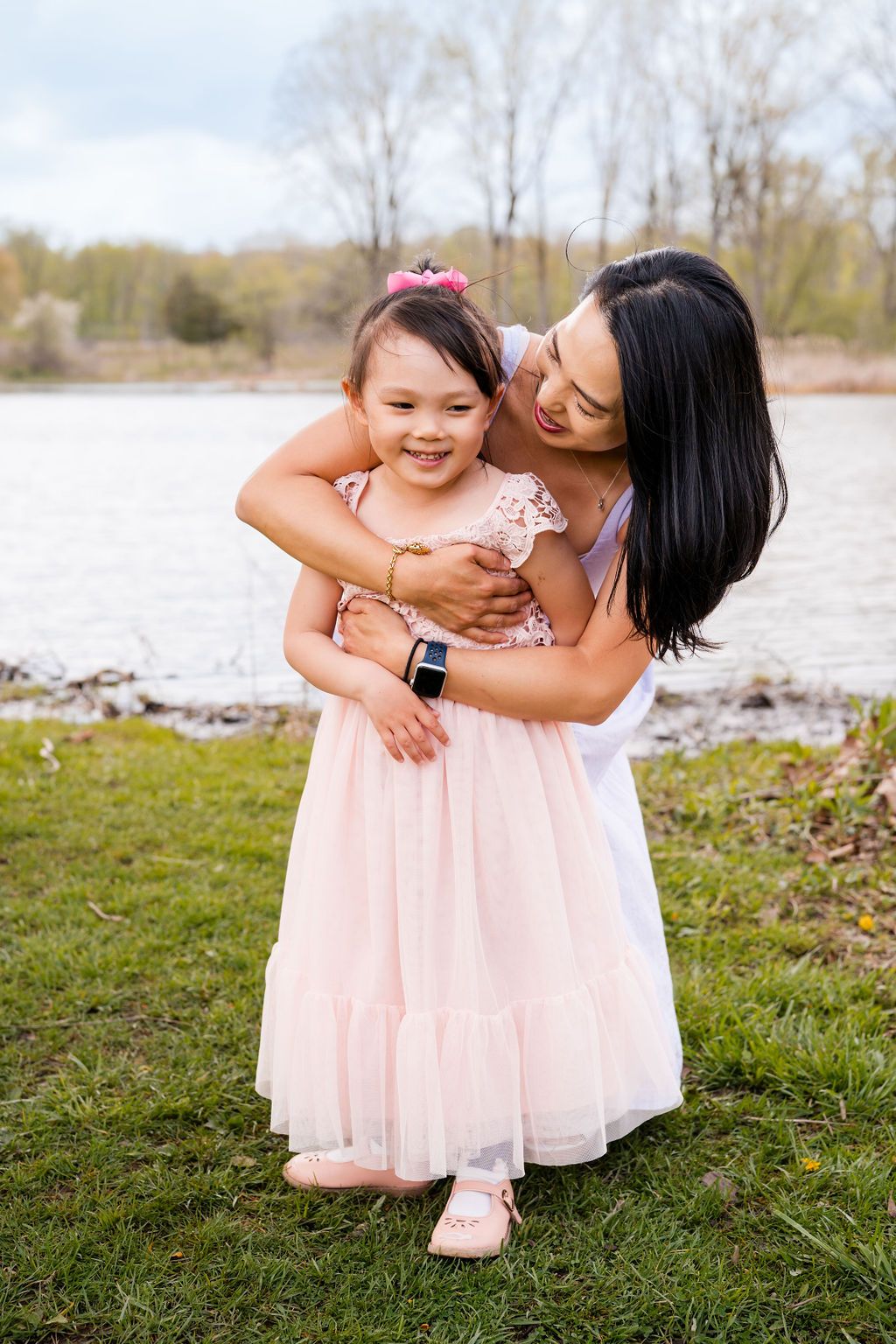 Mother and daughter share tender moments in pink dresses outdoors by the water.