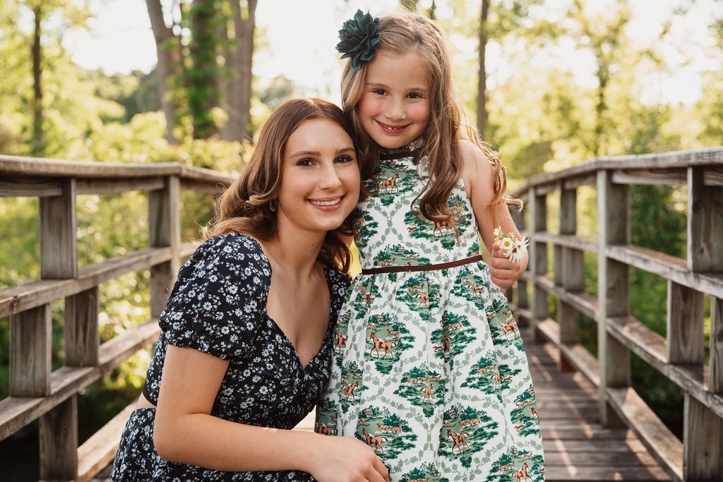 People in coordinating floral print dresses pose together on a wooden bridge in an outdoor setting.