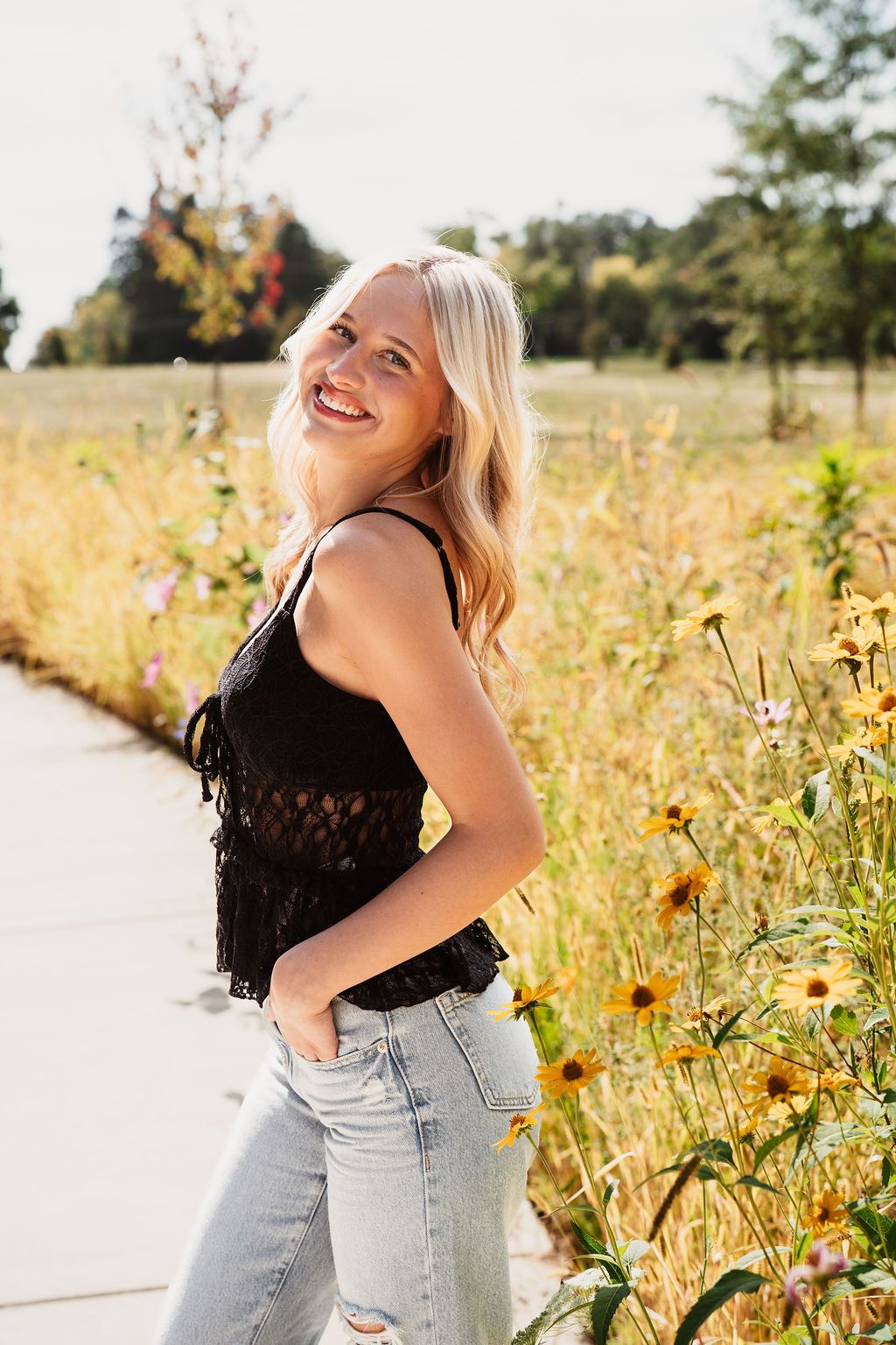 A smiling blonde woman in a black top and jeans poses beside wildflowers on a sunny day.