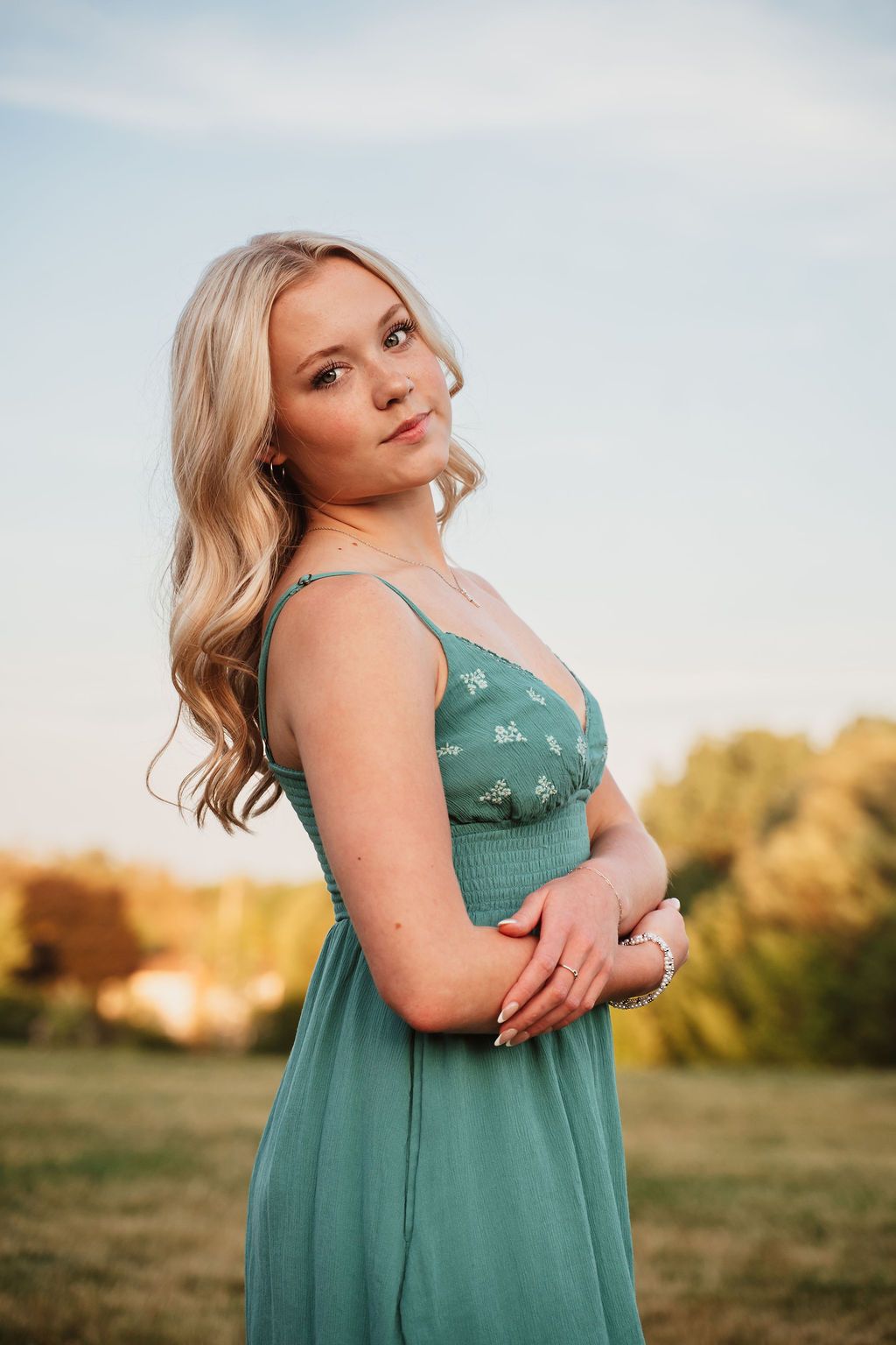 Young blonde woman in a teal floral sundress posing outdoors at golden hour in a green field.