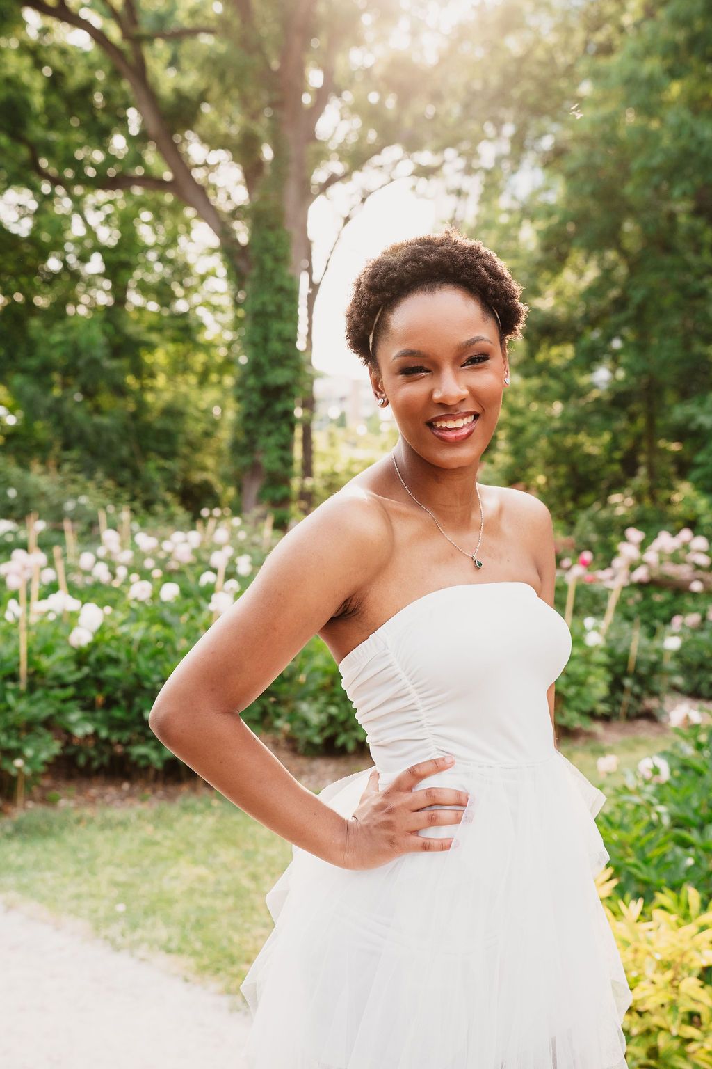A beautiful bride in a white strapless wedding dress poses outdoors in a sunlit garden setting with flowers.