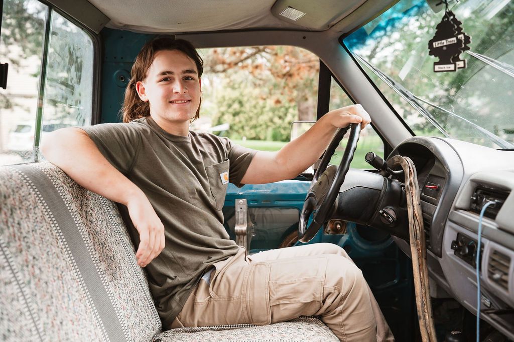 Someone sits in the driver's seat of a car with patterned upholstery and vintage interior details.