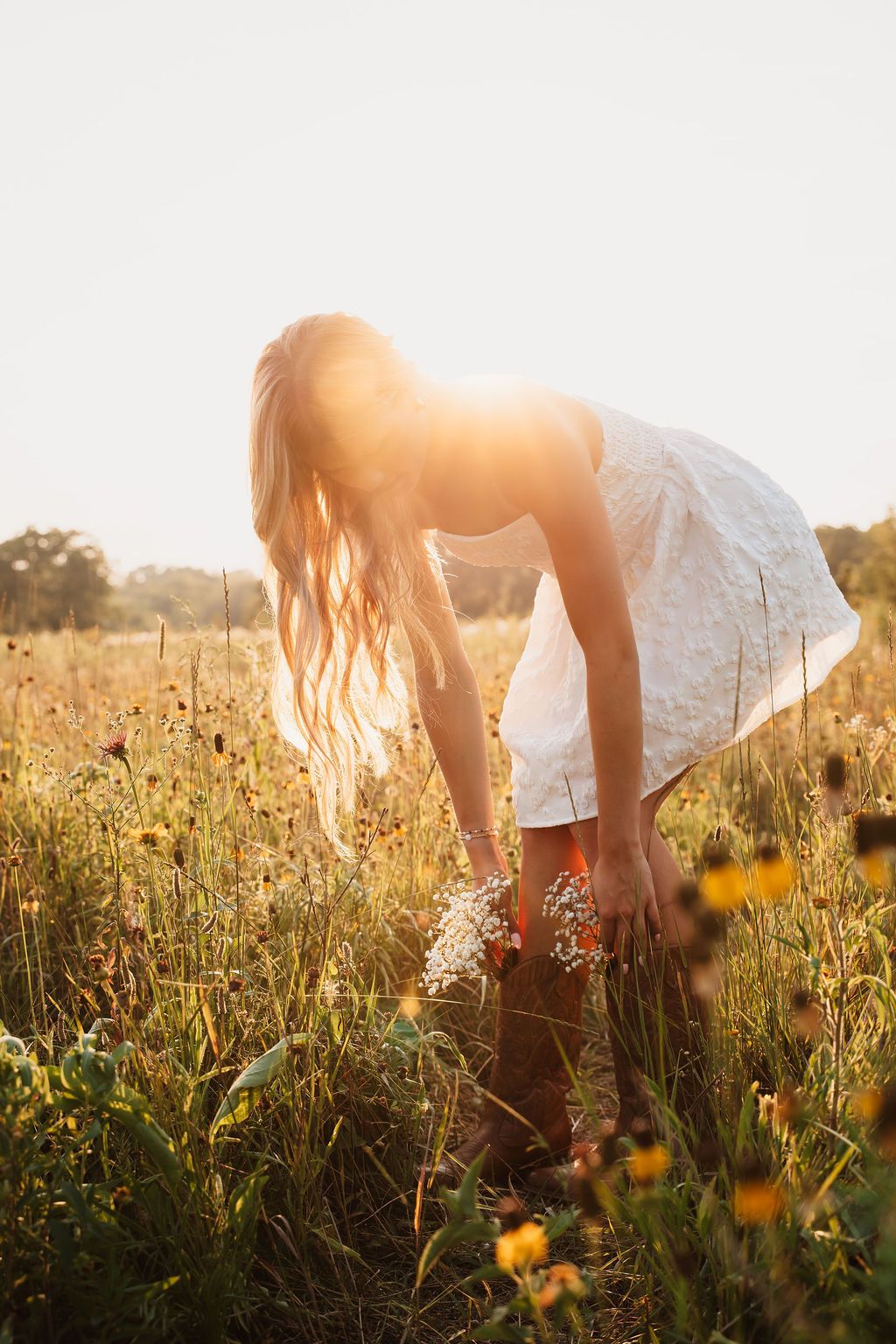 Woman in white dress picking wildflowers in a sunlit golden meadow at sunset.