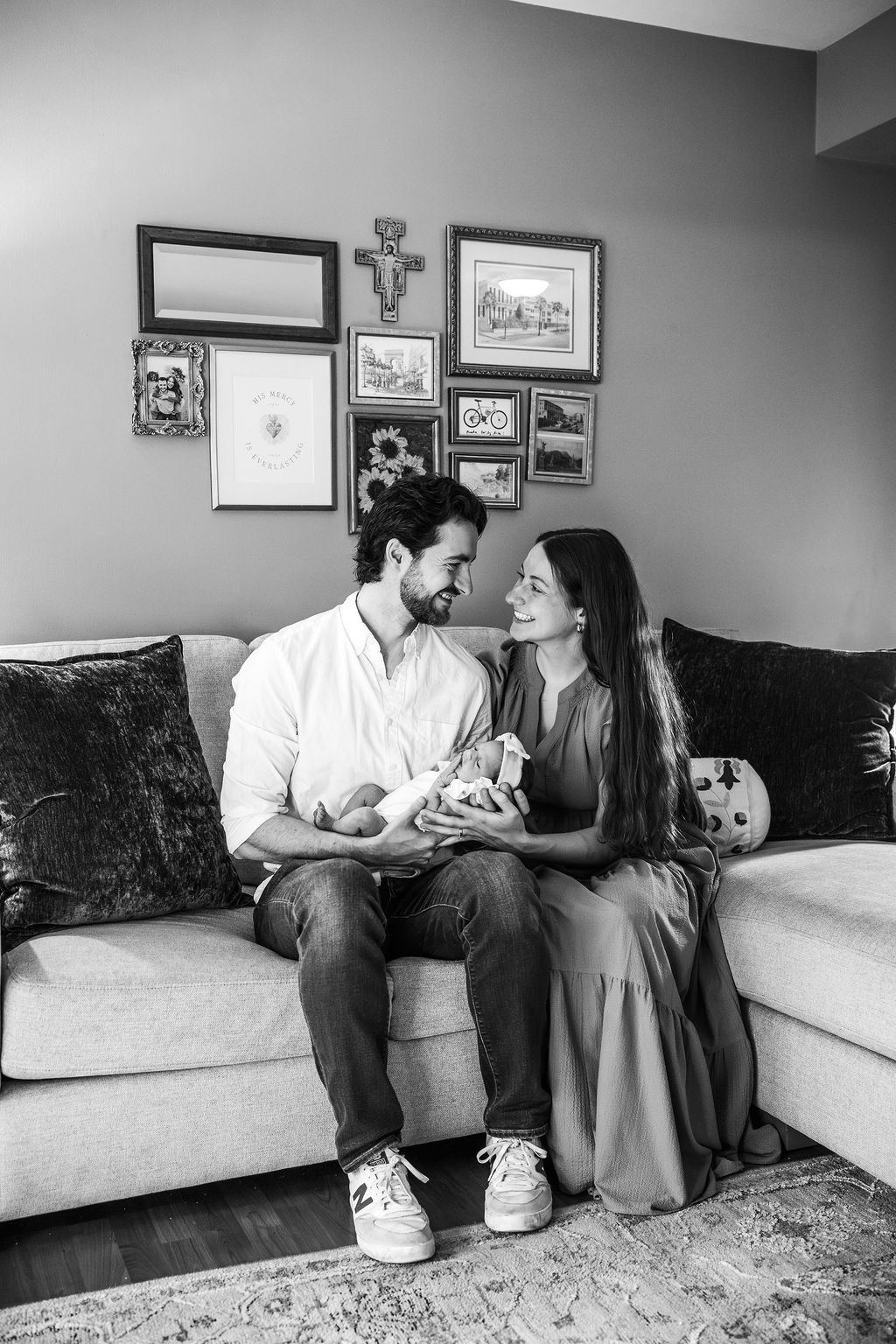 Black and white photo of a couple sitting on a couch, smiling at each other while holding their newborn baby.