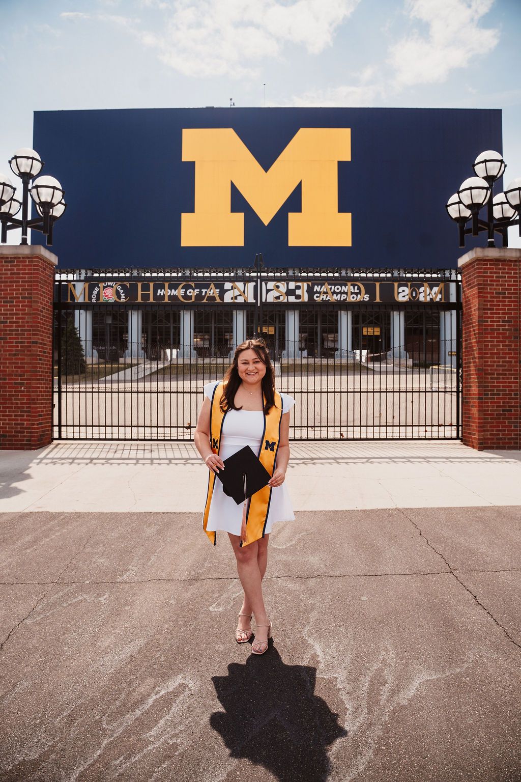 University of Michigan graduate poses in cap and gown outside the iconic Michigan Stadium gate with the block M logo.