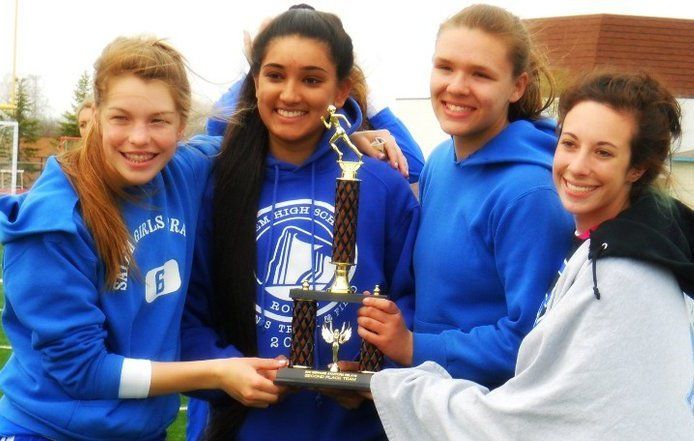 Four girls in blue hoodies proudly holding a trophy at an outdoor sports event.