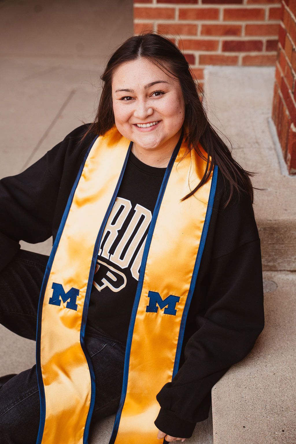 Smiling graduate wearing gold Michigan stole over black Purdue shirt, seated outdoors near brick wall.